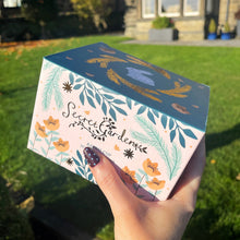 Hand holding a box labeled 'Secret Garden' with decorative elements against a garden background
