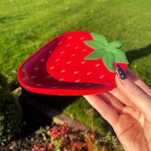 Hand holding a red strawberry-shaped object with green leaves against a grassy outdoor background