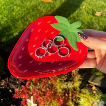 Red strawberry-shaped case with rings held by a hand outdoors.