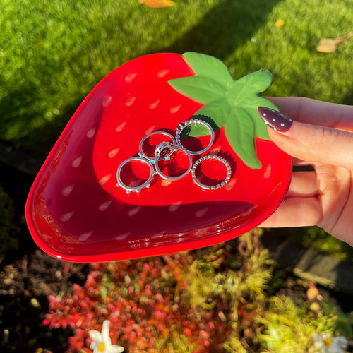 Red strawberry-shaped case with rings held by a hand outdoors.