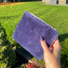 Hand holding a purple suede pouch outdoors with green grass and a building in the background
