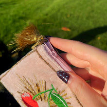 Hand holding a pink embroidered pouch with floral designs against a green grass background