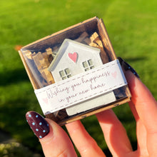 Hand holding a small box with a house-shaped card and a heart, outdoors on grass.