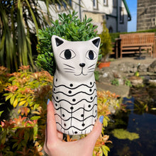 Cat-shaped planter with green plant held by a hand in a garden setting