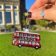 Red London bus ornament held by a hand with green nail polish against a blurred outdoor background.