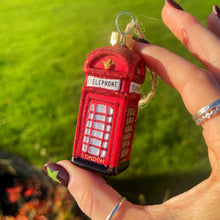 Hand holding a red telephone box ornament with a garden in the background