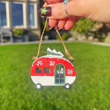 Hand holding a red and white Christmas ornament shaped like a camper on a grassy background