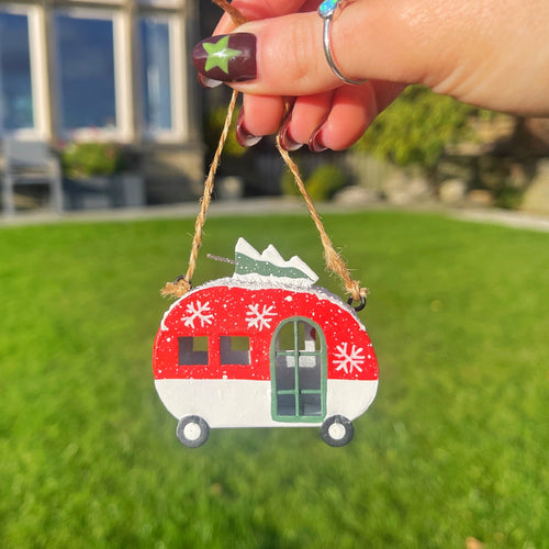 Hand holding a red and white Christmas ornament shaped like a camper on a grassy background