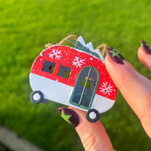 Hand holding a red and white camper-shaped ornament with snowflakes against a grassy background