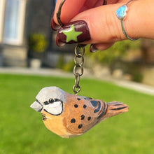 Hand holding a keychain with a bird design against a blurred outdoor background