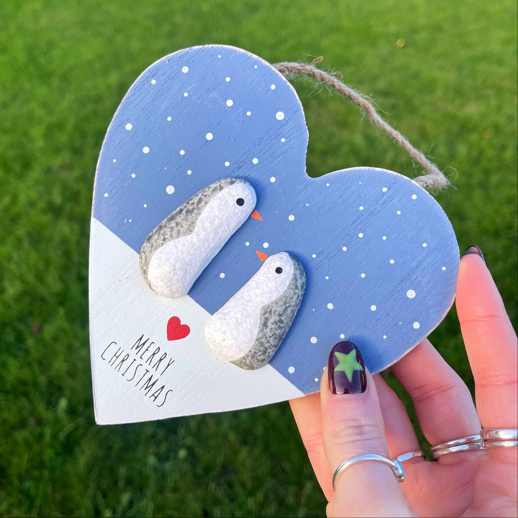 Heart-shaped wooden ornament with two birds and 'Merry Christmas' text, held against a grass background.
