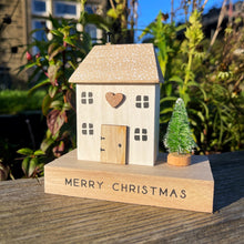 Wooden house ornament with a heart and Christmas tree on a 'Merry Christmas' block, outdoors.