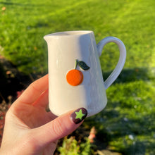 Small white pitcher with orange and green design held in front of a grassy outdoor background