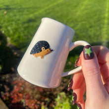 White mug with a hedgehog design held by a hand against a green outdoor background